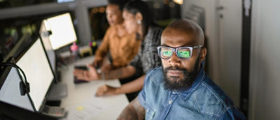 A man with glasses and a beard sitting at a table with others in the background.