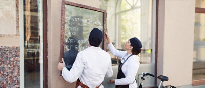 Dve osobe zaposlene u restoranu u belim uniformama i sa crnim šeširima nameštaju veliki meni na tabli ispred restorana