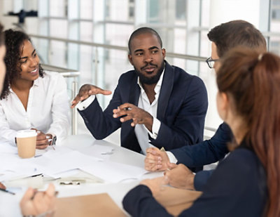 A group of five individuals is engaged in a conversation around a table in a bright office setting. 