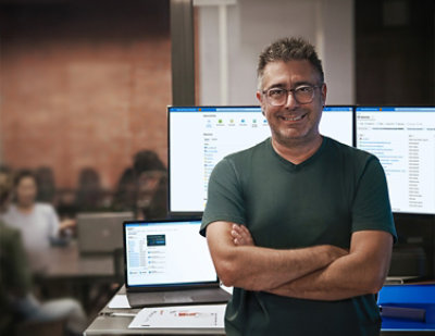 A man wearing a green shirt stands with his arms crossed in front of a computer.
