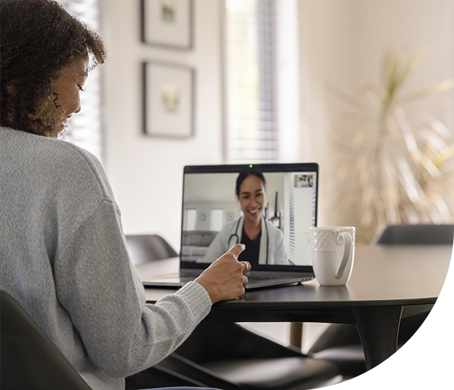 Woman wearing a light sweater is seated at a table in a bright room, engaged in a video call on a laptop. 