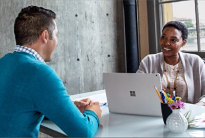 Two individuals in a meeting with one person using a laptop and the other taking notes, sitting at a table with a vase of flowers.