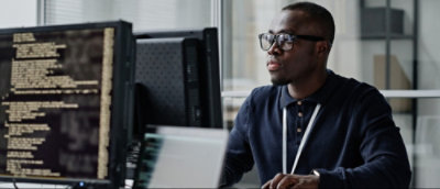 A person wearing glasses is sitting at a desk, looking at a computer screen displaying code, in an office setting.