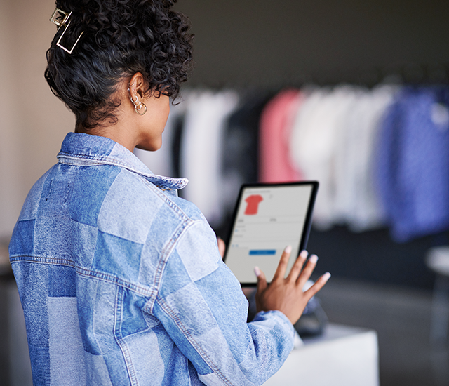 A person with curly hair and wearing hoop earrings, dressed in a patchwork denim jacket, interacts with a tablet displaying a red shirt. They are in a clothing store, with blurred racks of various garments, including white, pink, and blue clothing, in the background. The scene suggests a digital shopping or inventory process within a retail environment.
