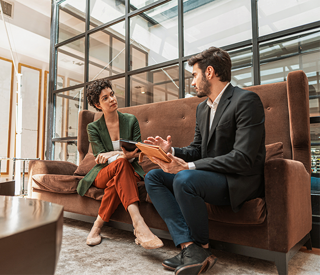 A man in a black suit and a woman in a green blazer and orange pants sit on a brown couch, engaged in conversation. The setting is a modern office with large glass windows and abundant natural light. Both hold notebooks or documents, suggesting a focused, professional discussion. The atmosphere is casual yet collaborative..