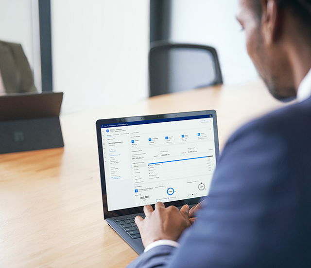 A person in a suit sits at a conference table, using a laptop with financial software on its screen. The software interface features various financial data and charts. In the background, another individual is partially visible, sitting with a closed laptop. The scene suggests a business meeting or financial planning session, highlighted by the conference room setting and professional attire.