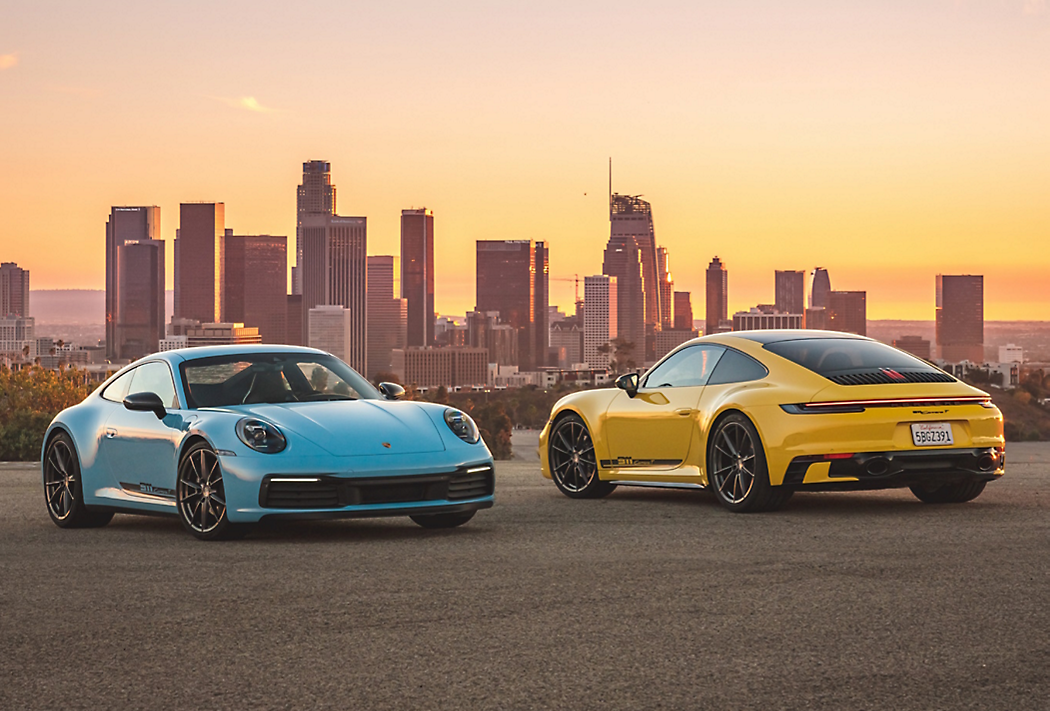 Dos coches Porsche, uno azul y otro amarillo, estacionados con un horizonte urbano en el fondo durante el atardecer.