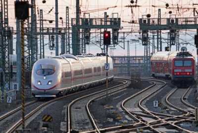 A modern high-speed train arriving at a station with multiple railway tracks and overhead wires at dusk.