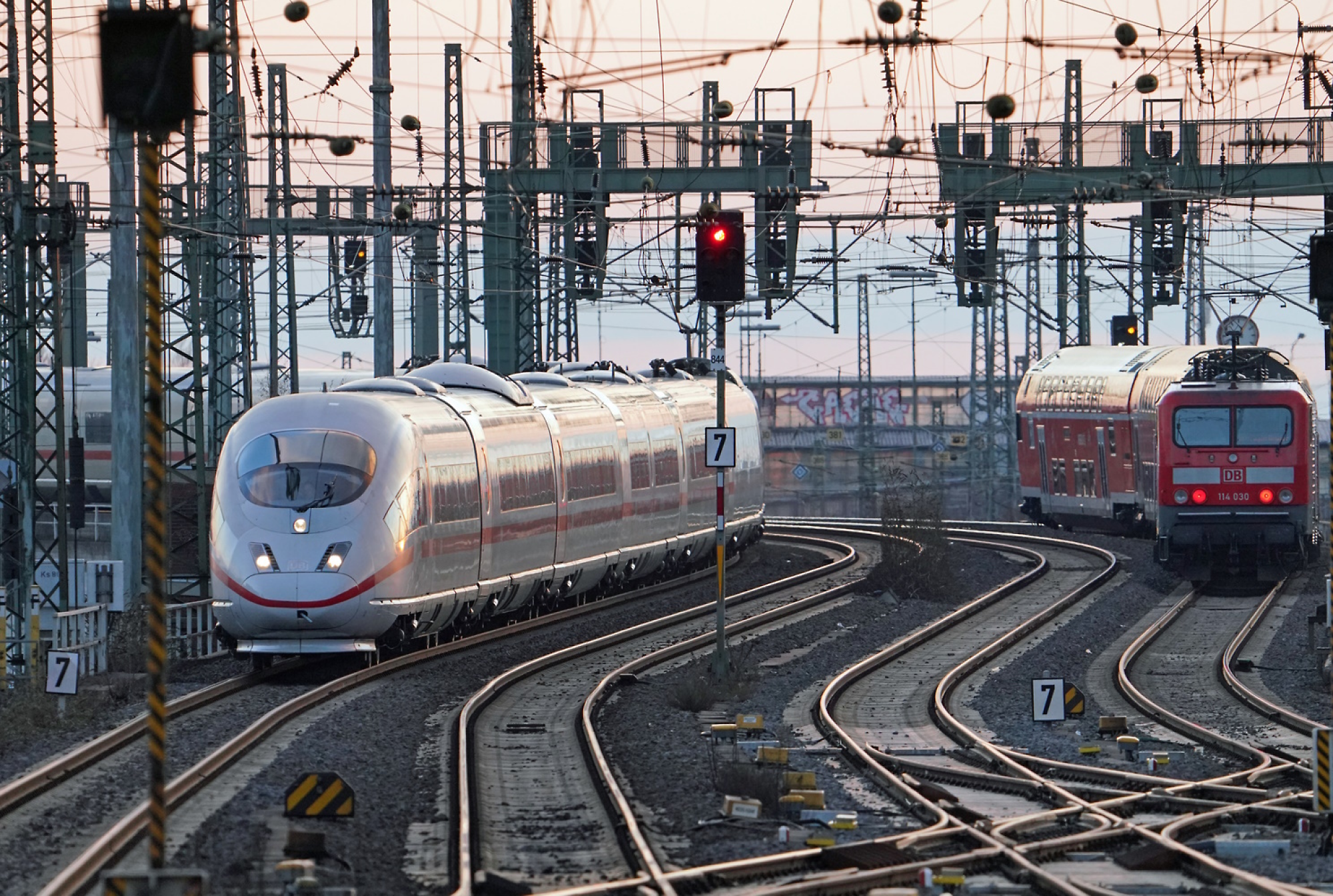 Un train moderne à grande vitesse arrivant dans une gare avec plusieurs voies ferrées et des câbles aériens au crépuscule.