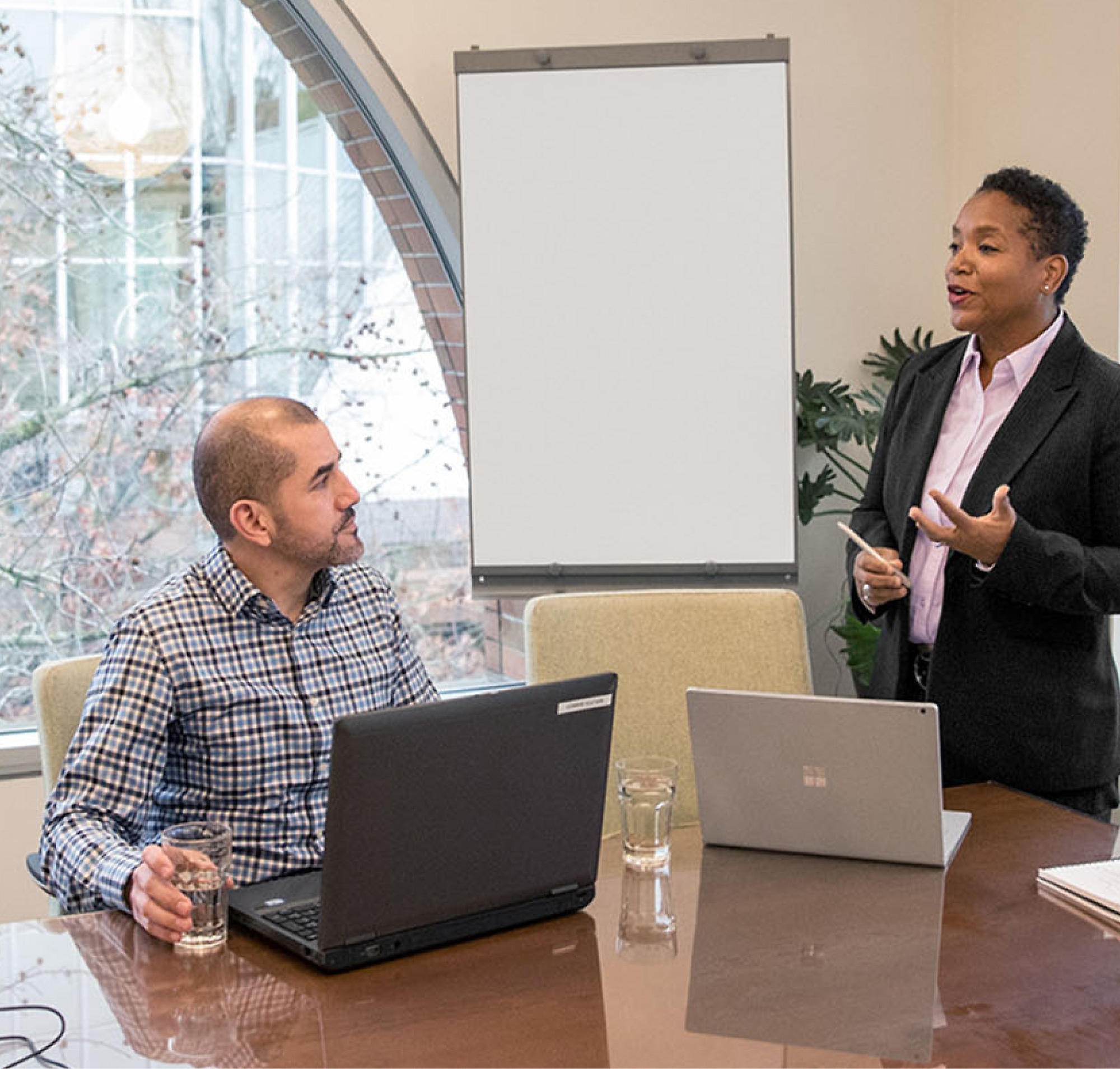 Two professionals in a meeting room one standing presenting and one seated at a table with laptops in front of a whiteboard.