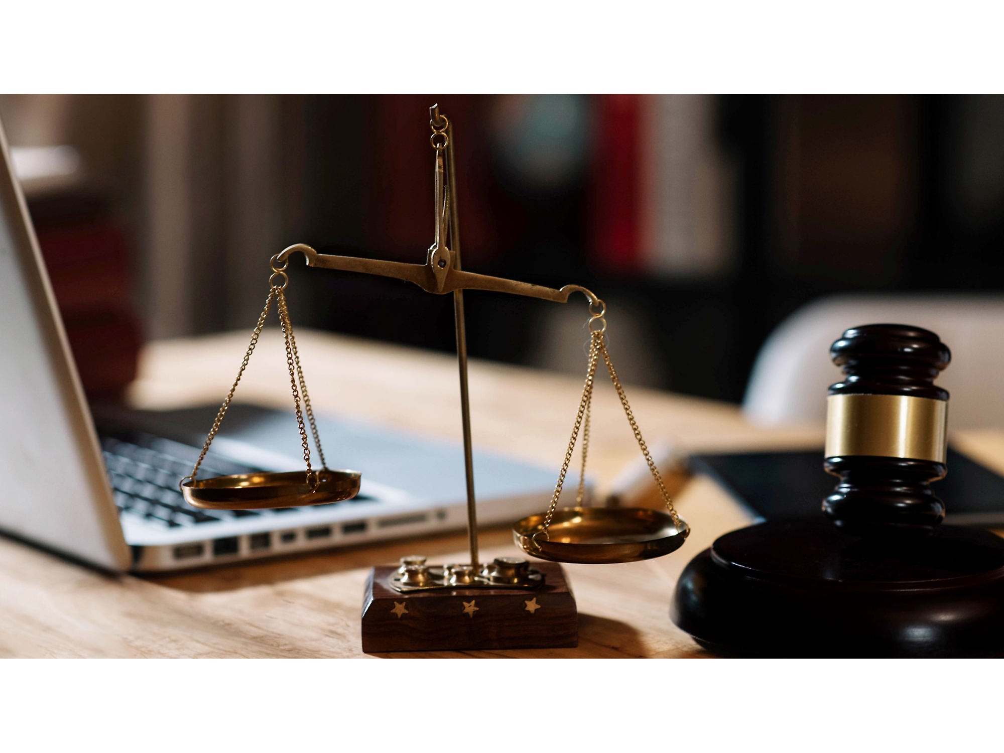 Puidust kohtunikuhaamer ja kaalude komplekt laual, taustal sülearvuti, mis sümboliseerib juriidilist ametialast tegevust kontoris.