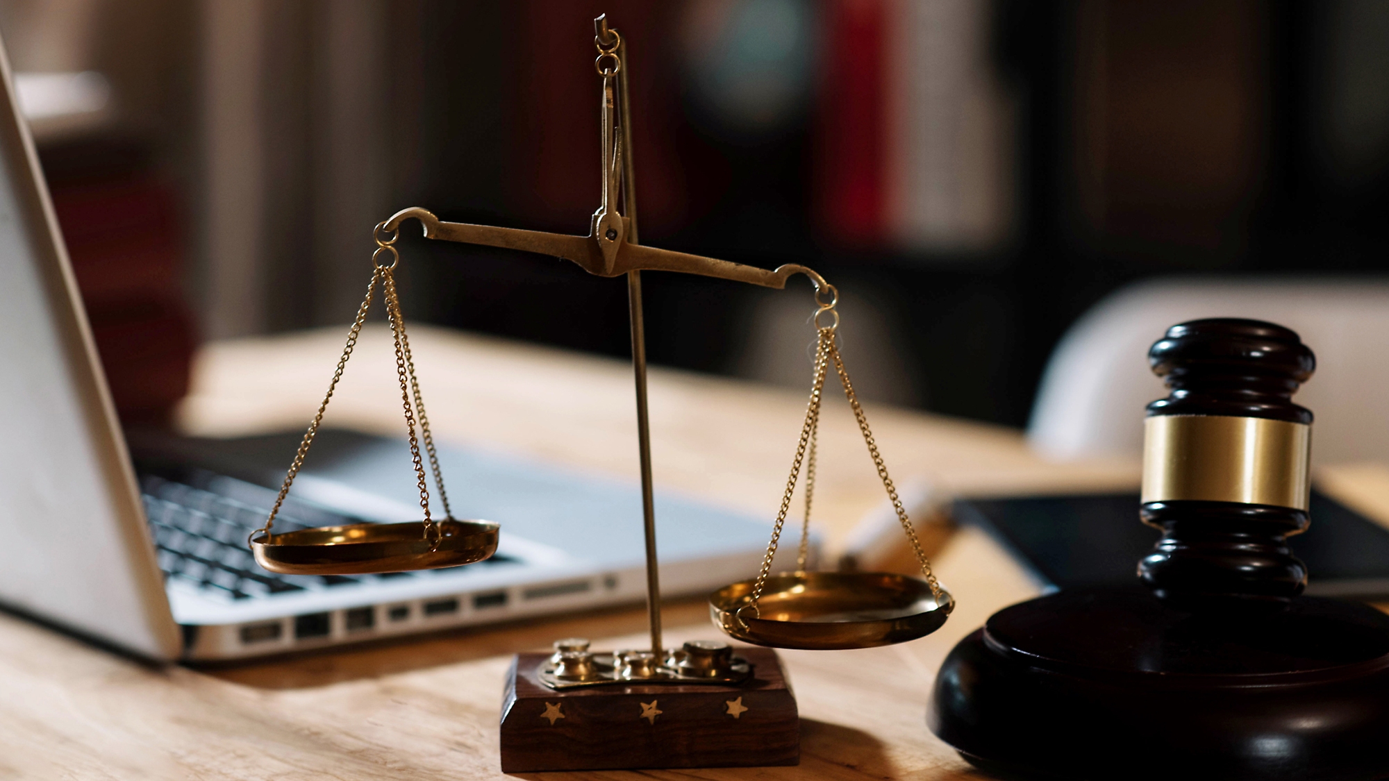 A wooden gavel and a set of scales on a desk, with a laptop in the background, symbolizing legal practice in an office setting.