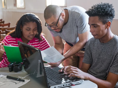Troje ljudi sarađuje na projektu koristeći laptop i dokumente za stolom u jako osvetljenoj prostoriji.