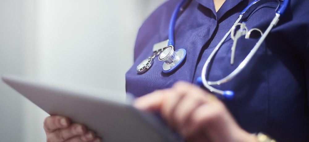 Doctor într-o uniformă medicală albastră ținând în mână o tabletă, cu un stetoscop atârnat la gât, verificând fișe medicale.