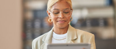 A professional woman with glasses and a light beige blazer reads something on her tablet in a modern office setting.