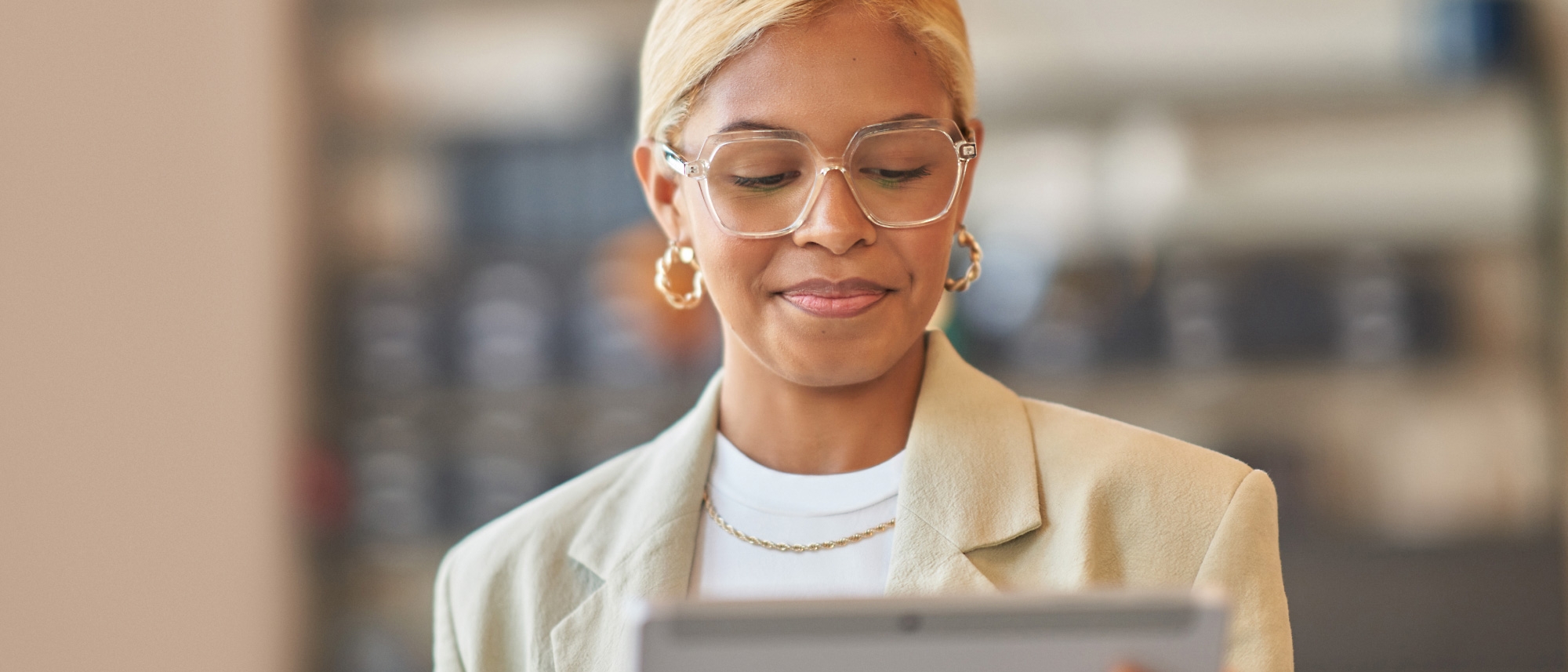 Una mujer profesional con gafas y una chaqueta de color marrón claro lee algo en su tableta en un entorno de oficina moderna.