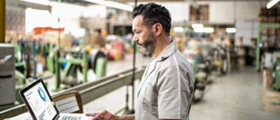 Sredovečni čovek sa sedom kosom i bradom, nosi svetlo sivu košulju i pregleda podatke na laptopu u industrijskom skladištu.
