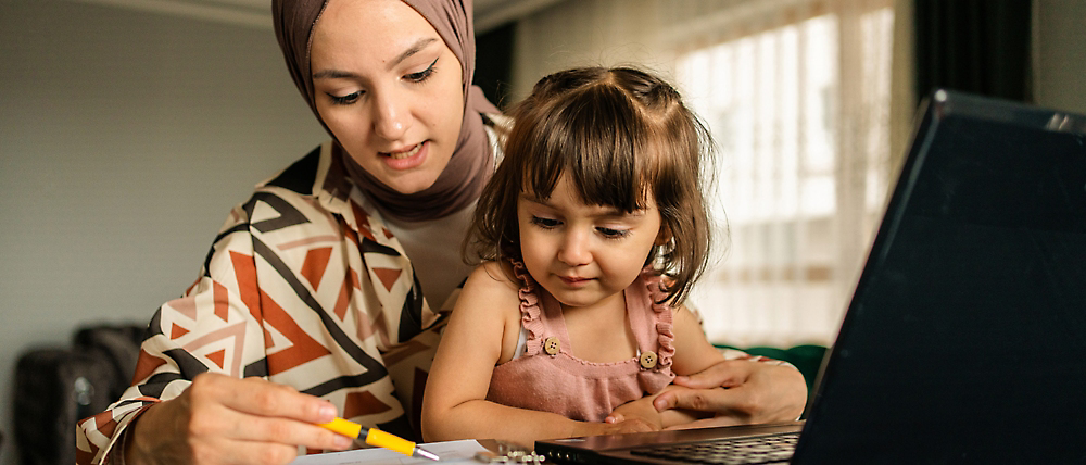 A woman wearing a hijab and a young child sitting at a desk, looking at a laptop screen together.