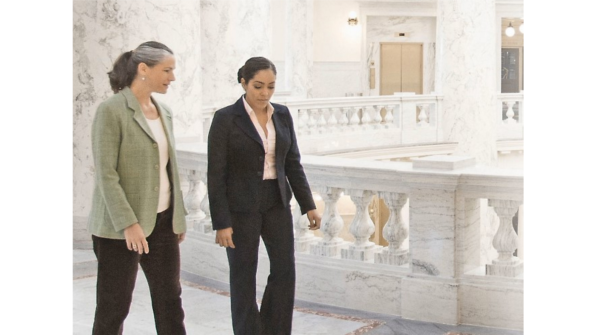 Two women walking and conversing in a marble hall with ornate balustrades in the background.