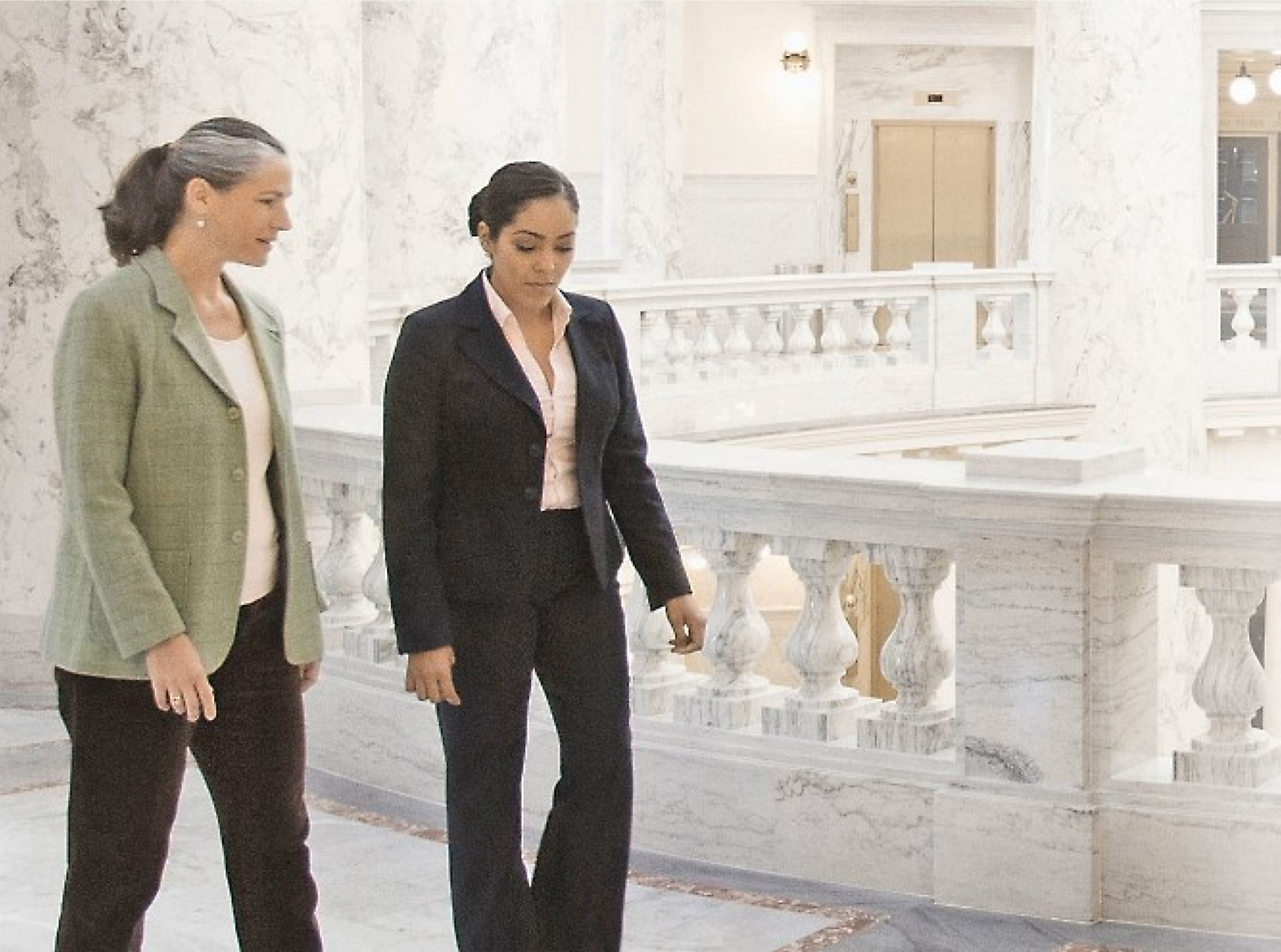 Two women walking and conversing in a marble hall with ornate balustrades in the background.