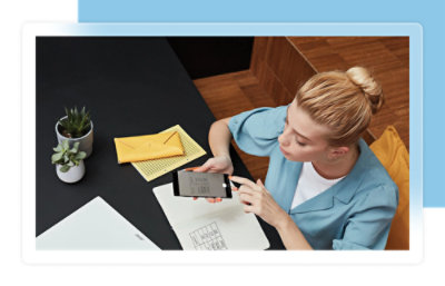 Une femme en chemise bleue utilisant un smartphone assise à une table avec un carnet de notes, une enveloppe jaune et des plantes en pot.
