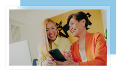 Deux femmes souriantes regardant l’écran d’un smartphone dans une pièce lumineuse, avec une œuvre d’art abstraite en arrière-plan.
