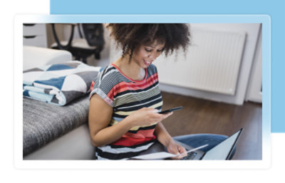 Une femme assise par terre, regardant son téléphone, avec un ordinateur portable et une couverture pliée à proximité.