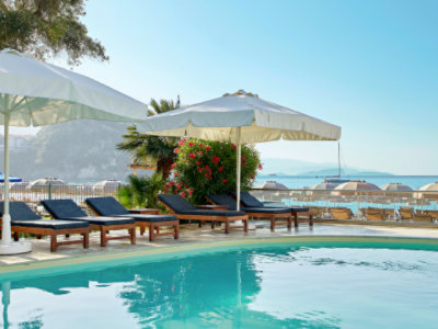 A poolside area with sun loungers and large umbrellas overlooks a beach with numerous umbrellas and sunbeds.