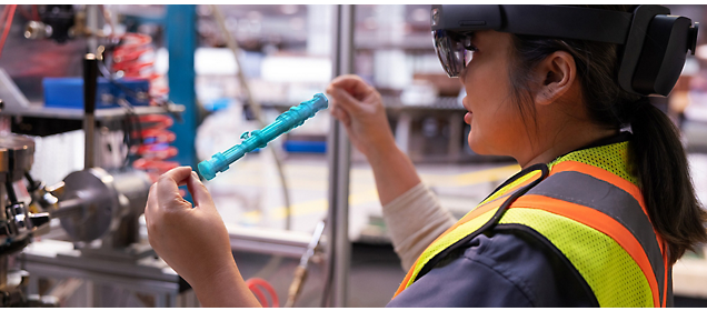 Um trabalhador usando um colete de segurança e óculos de realidade aumentada inspeciona um componente industrial.