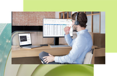 在现代化办公室里，一位头戴耳机的男士坐在办公桌前，使用笔记本电脑和台式机，其中的电子表格已打开。