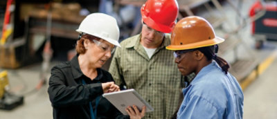 Three construction workers with hard hats discussing over a tablet in a warehouse.