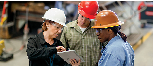Três trabalhadores da construção civil com capacetes discutindo sobre um tablet em um armazém.