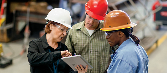 Three construction workers with hard hats discussing over a tablet in a warehouse.