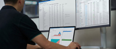 A person working on a laptop with data analysis graphs on the screen, in front of a larger monitor 