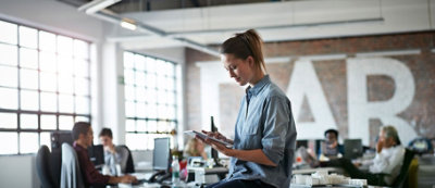 Una donna in piedi in primo piano usa il proprio telefono, mentre molte persone sono sedute e lavorano al computer in un ufficio tipo open-space 