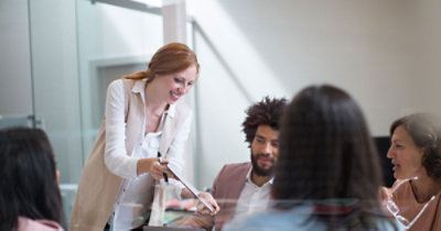 Žena pruža dokument muškarcu sa laptop računarom u sobi za sastanke, dok još tri osobe
