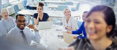 Różnorodna grupa ludzi siedzi przy stole podczas spotkania, niektórzy z nich z laptopami. Osoba na pierwszym planie wydaje się przemawiać lub prezentować grupie.