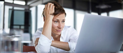 אדם בשיער קצר, לובש חולצה לבנה, יושב ליד שולחן ומביט בלחץ תוך שימוש במחשב נישא.