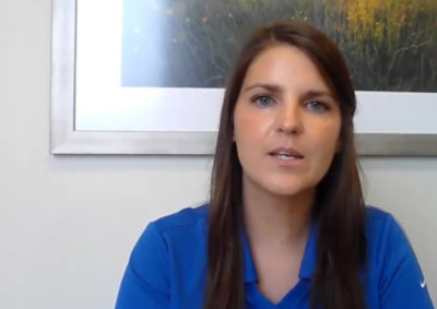 Woman with long brown hair wearing a blue shirt sits in front of a framed picture, looking forward.