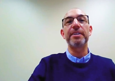 A person with glasses and a beard wearing a dark sweater and blue shirt, seated against a plain light-colored background.