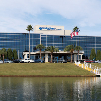 A large, modern glass-fronted building labeled "Singing River Hospital" with palm trees and a U.S. flag in front, across a small body of water.