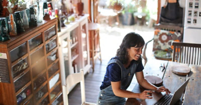Žena sedi za drvenim stolom i koristi laptop u prostoriji sa drvenim nameštajem i raznim predmetima za domaćinstvo.
