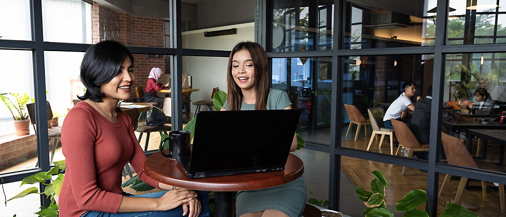 Two women discussing and working on a laptop at a round table in a busy café with large windows and plants.