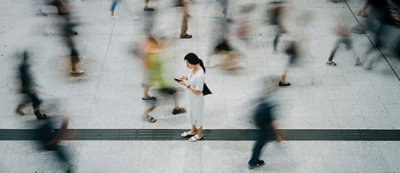 אישה עומדת מבלי לזוז, מביטה בטלפון שלה בשטח ציבורי עמוס, בעוד שאנשים סביבה נמצאים בתנועה, ויוצרים אפקט מטושטש.