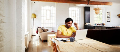 Osoba w żółtej koszuli siedzi przy drewnianym stole, korzystając z laptopa w jasnym, przestronnym salonie z dużymi oknami 