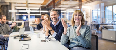 Grupa ludzi siedzi przy stole konferencyjnym w biurze, bijąc brawo i uśmiechając się. Na stole leżą laptopy, dokumenty i kubek.