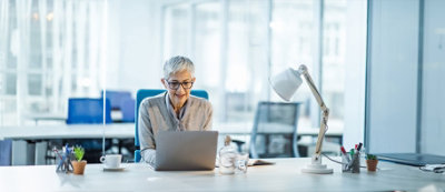 Osoba z krótkimi siwymi włosami i w okularach siedzi przy biurku w biurze, pracując na laptopie.