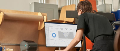 A person in a black shirt leans over a desk, interacting with data on a large desktop screen showing graphs and a map