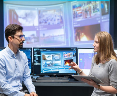 A man and woman sitting in front of multiple computer screens, with the woman holding a tablet.