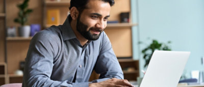 A person sitting at a desk, looking at a laptop screen, with shelves containing books and plants in the background.